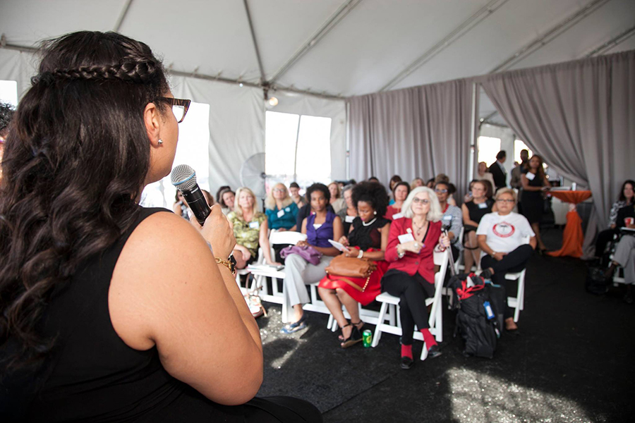 A diverse group of women in an audience listening to a woman standing in front of them talk